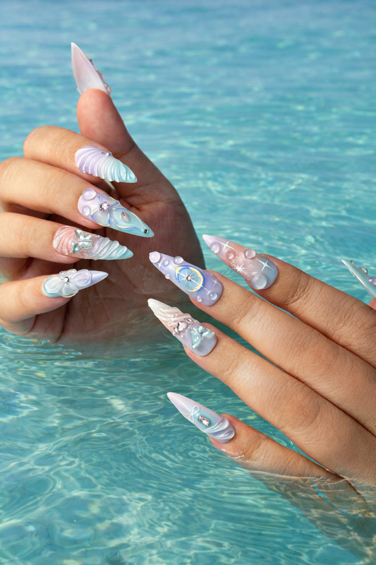 Pastel press-on nails with 3D seashell and pearl designs on a hand gently touching clear turquoise ocean water, creating a soft and refreshing summer vibe.